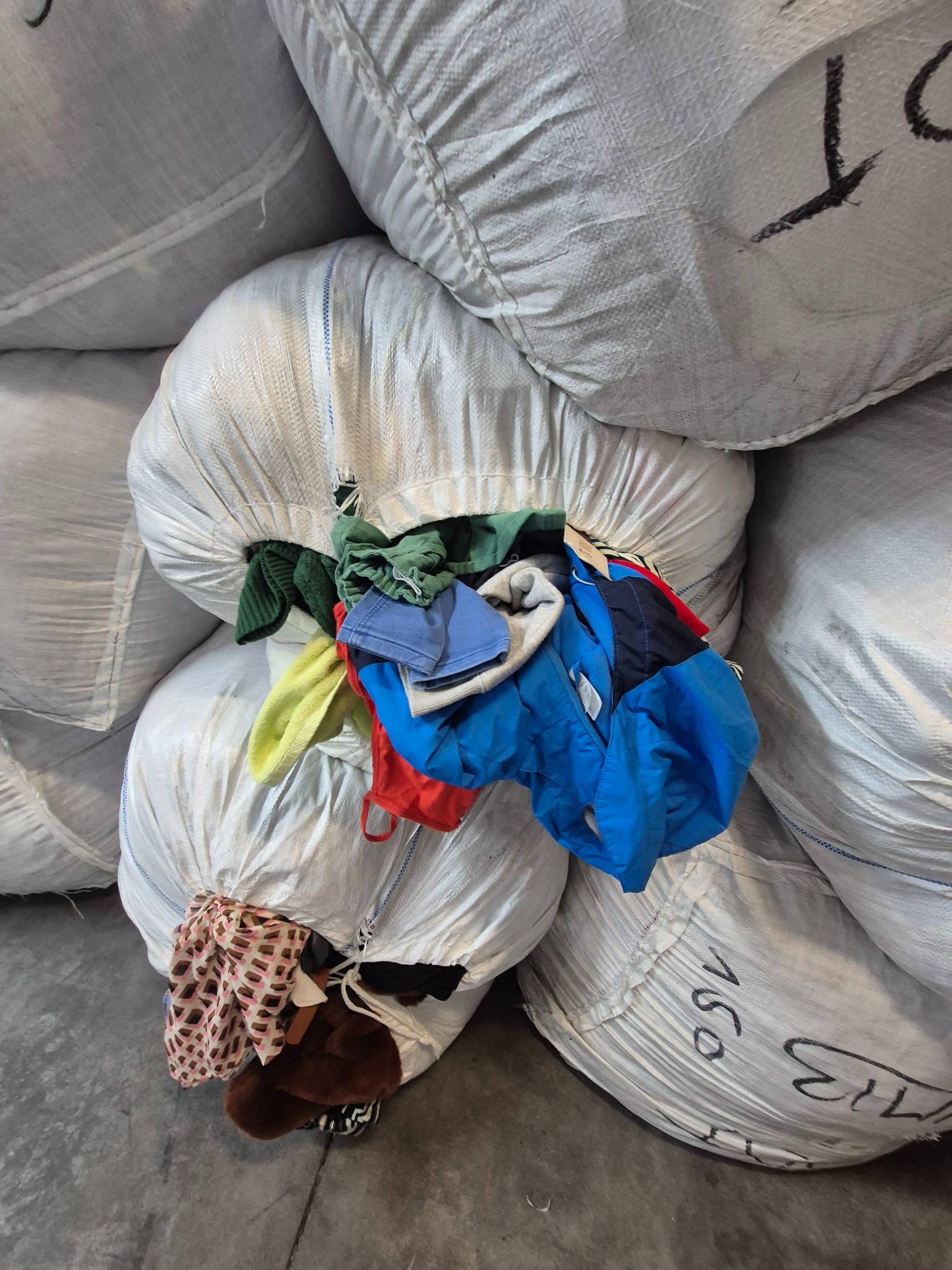 Stos worków z odzieżą używaną, na wierzchu widoczne ubrania w różnych kolorach i fasonach. Worki z szarego materiału z czarnymi napisami, ustawione jeden na drugim.