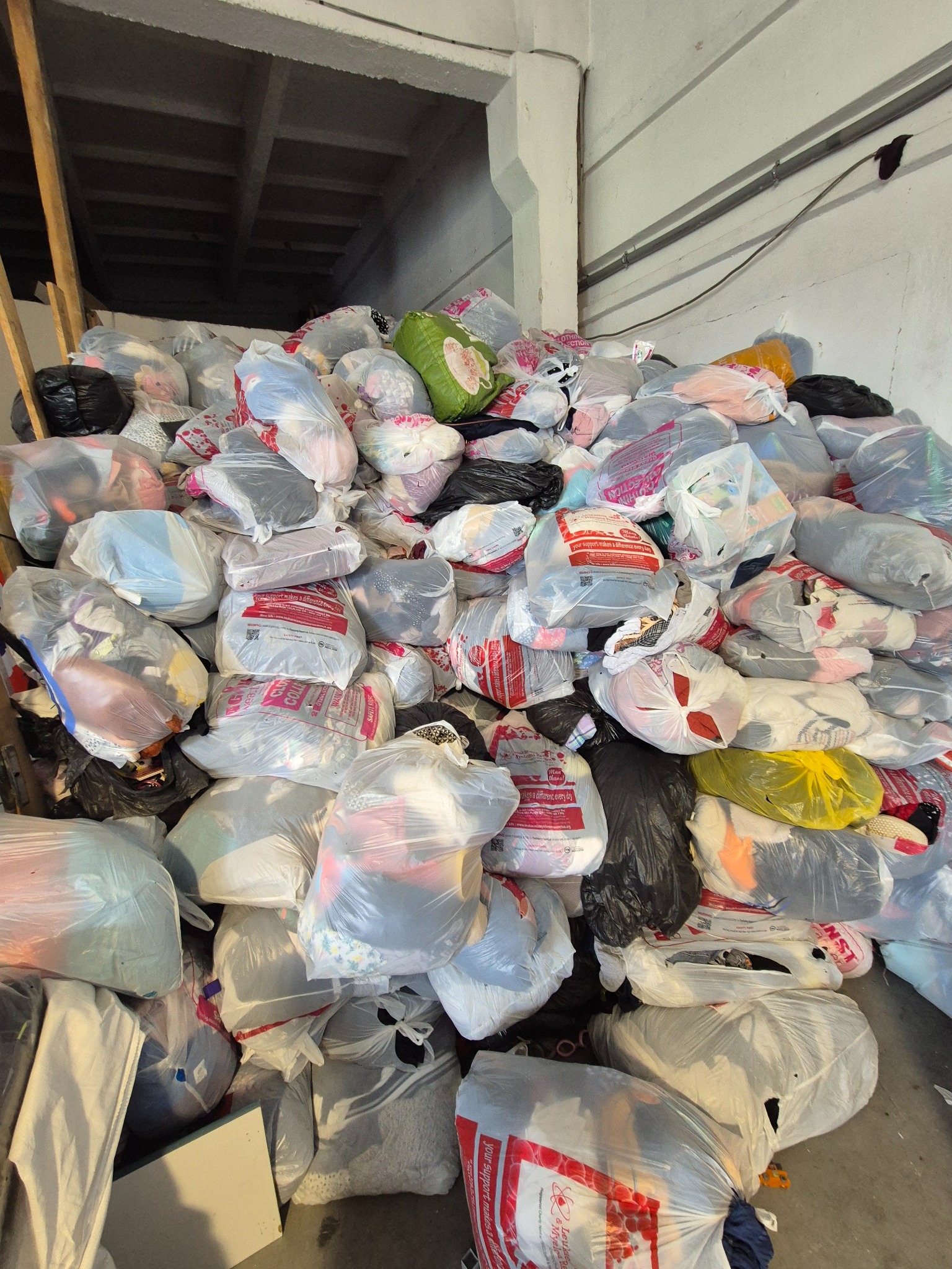 Góra worków z odzieżą używaną w magazynie, widoczne etykiety i różne kolory ubrań. Częściowo widoczne elementy konstrukcyjne budynku.