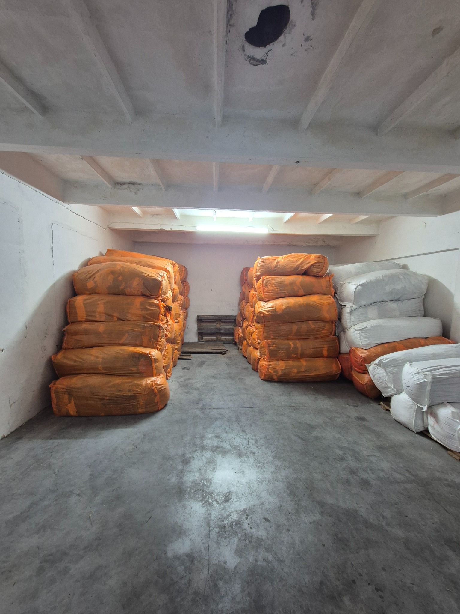 Magazyn z workami odzieży używanej w pomarańczowych i białych workach, ułożonymi w stosy na betonowej podłodze w pomieszczeniu z betonowym sufitem.