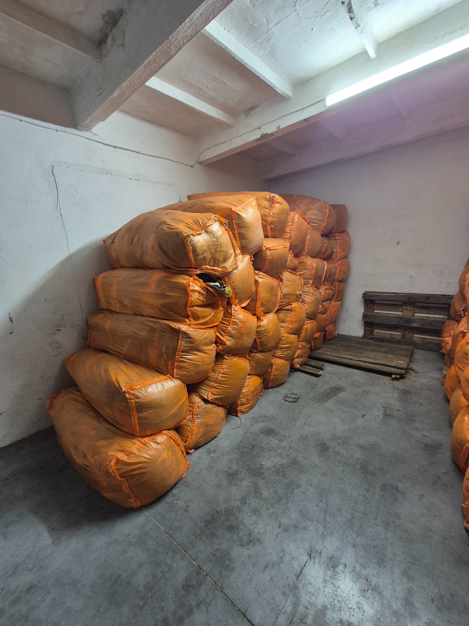 Pomarańczowe worki ułożone w magazynie, niektóre z widoczną zawartością, palety w tle, surowy beton na podłodze i ścianach.