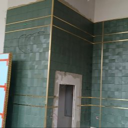 GALBUD SYLWIA GALOR - Ściana wyłożona zieloną, wzorzystą glazurą z dekoracyjnymi, złotymi listwami. Widoczny laser krzyżowy i przygotowane płyty do montażu. W tle rusztowania.