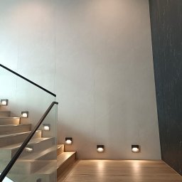 GALBUD SYLWIA GALOR - Nowoczesne schody z drewnianymi stopniami i szklaną balustradą, oświetlone punktowymi lampami ściennymi. Ściana wykończona jasnymi płytami imitującymi kamień.