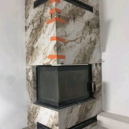 GALBUD SYLWIA GALOR - Obudowa kominka z płytek imitujących marmur w trakcie montażu. Widoczne pomarańczowe taśmy i poziomice. Fragment podłogi i ściany w tle.