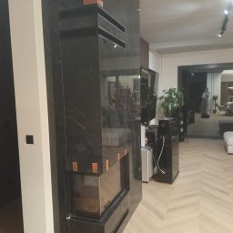 GALBUD SYLWIA GALOR - Elegancki kominek obłożony czarnym marmurem z widocznymi detalami wykończeniowymi i elementami montażowymi, w nowoczesnym salonie z parkietem w jodełkę.