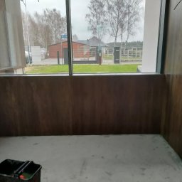GALBUD SYLWIA GALOR - Przygotowane do malowania wnętrze z brązowymi panelami ściennymi i widokiem na zewnątrz przez duże okno. Wiadro z wałkami malarskimi w rogu.
