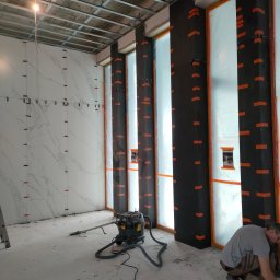 GALBUD SYLWIA GALOR - Wnętrze w trakcie prac glazurniczych: ściana z białych płytek imitujących marmur, czarne kolumny zabezpieczone folią, widoczny pracownik i odkurzacz przemysłowy.