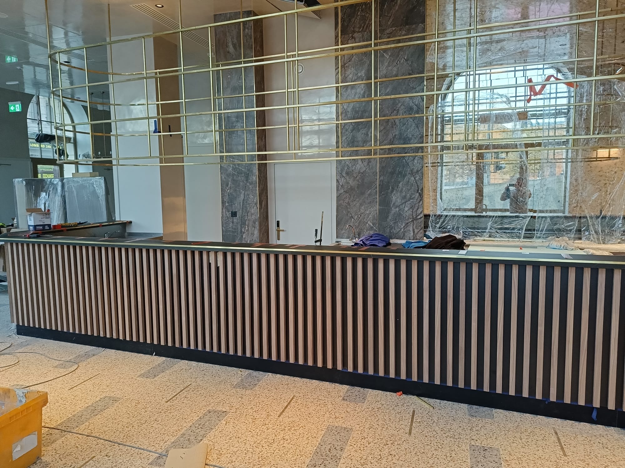 Nowoczesna zabudowa baru z pionowymi panelami w odcieniach drewna i czerni, złota konstrukcja nad blatem, marmurowe wykończenie ścian w tle. Wnętrze w trakcie wykończenia.