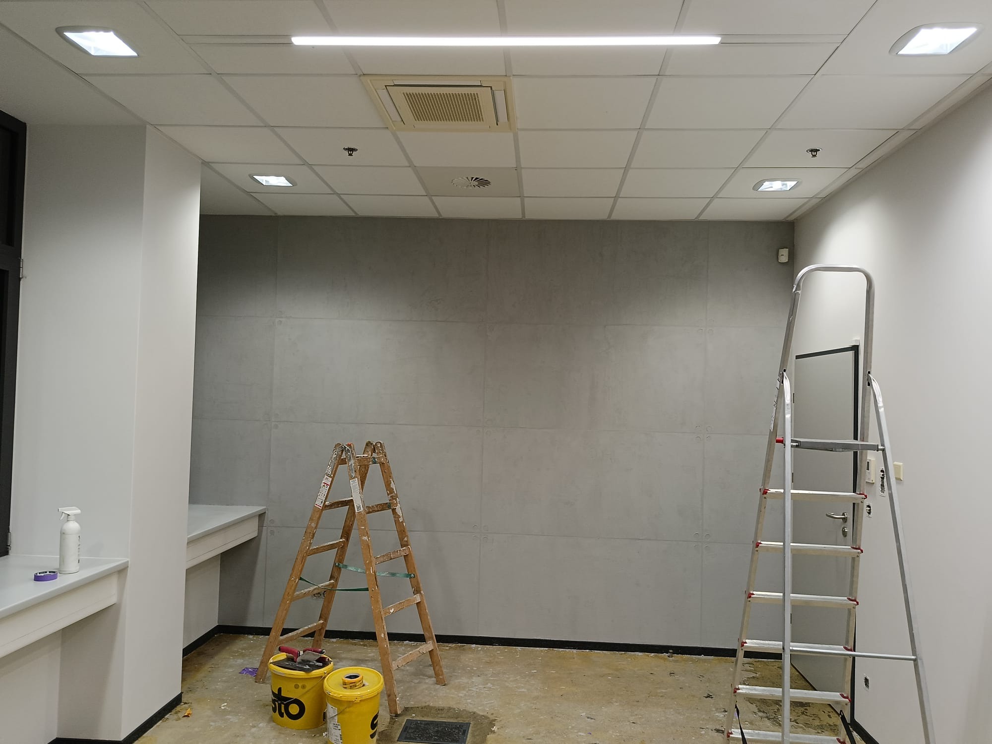 Wnętrze biura w trakcie prac wykończeniowych. Widoczne drabiny, wiaderka z farbą i przygotowana ściana. Oświetlenie sufitowe i minimalistyczny design.