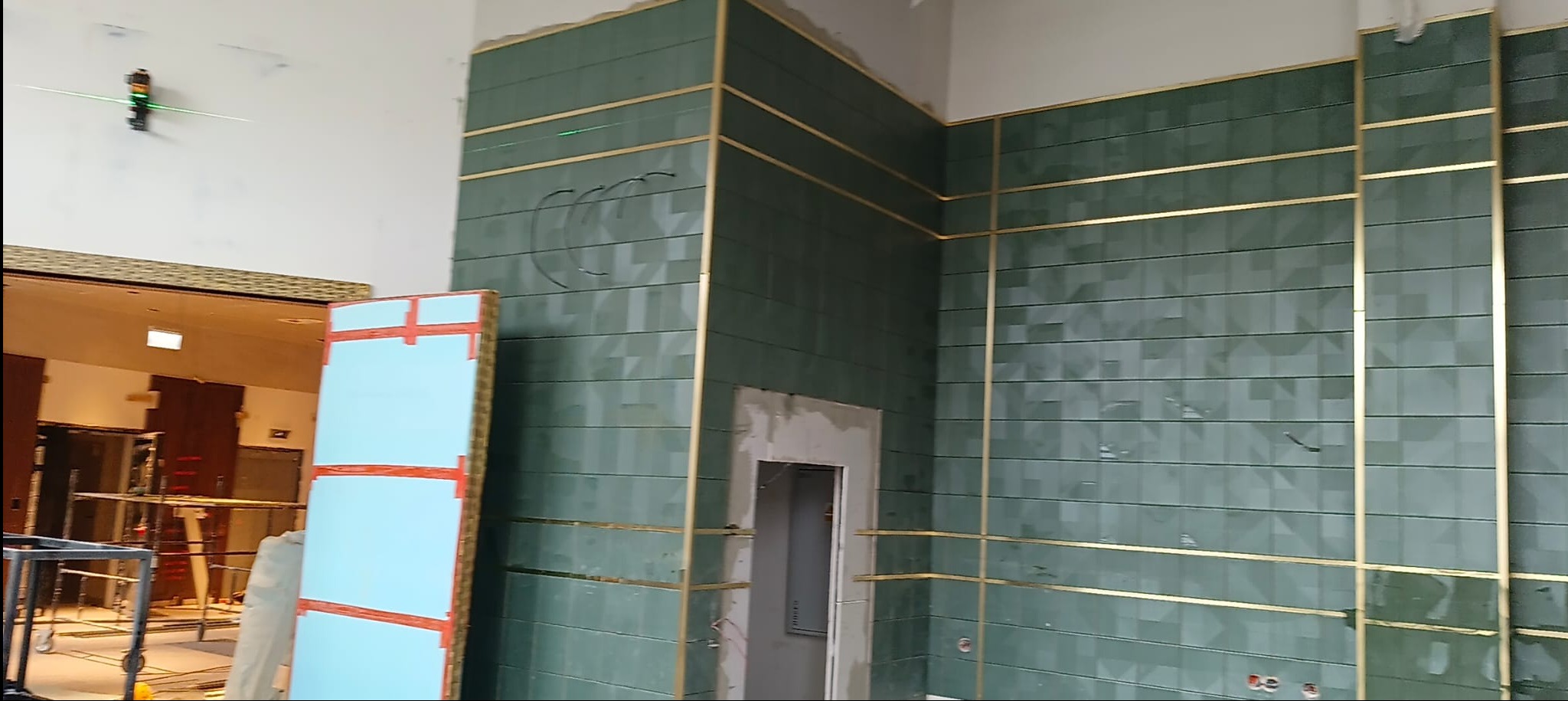 Ściana wyłożona zieloną, wzorzystą glazurą z dekoracyjnymi, złotymi listwami. Widoczny laser krzyżowy i przygotowane płyty do montażu. W tle rusztowania.