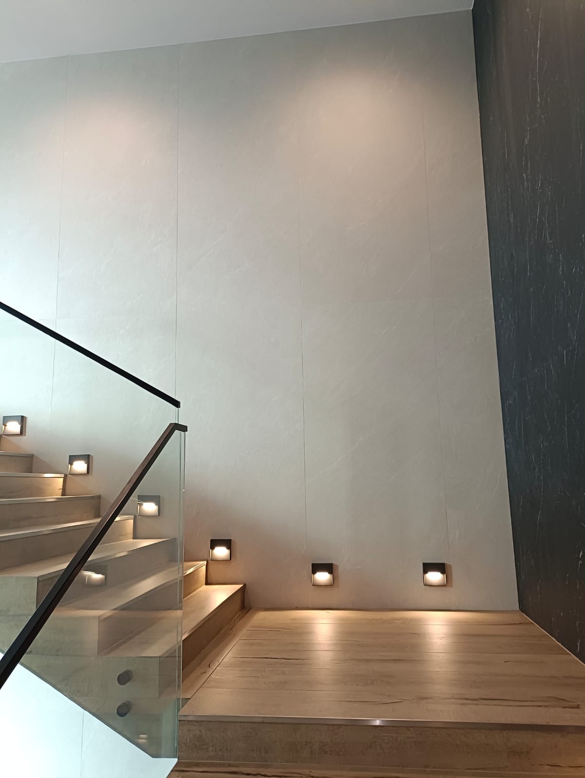 Nowoczesne schody z drewnianymi stopniami i szklaną balustradą, oświetlone punktowymi lampami ściennymi. Ściana wykończona jasnymi płytami imitującymi kamień.