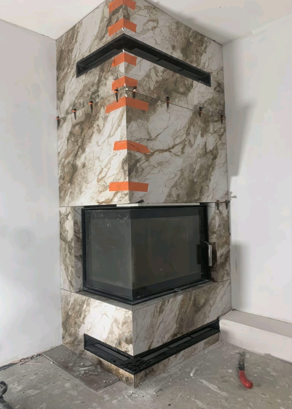 Obudowa kominka z płytek imitujących marmur w trakcie montażu. Widoczne pomarańczowe taśmy i poziomice. Fragment podłogi i ściany w tle.