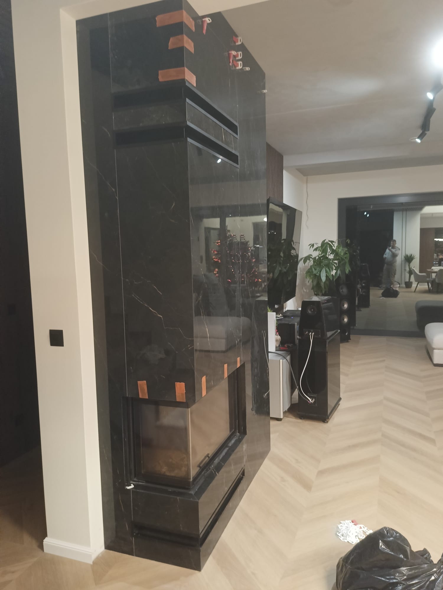 Elegancki kominek obłożony czarnym marmurem z widocznymi detalami wykończeniowymi i elementami montażowymi, w nowoczesnym salonie z parkietem w jodełkę.