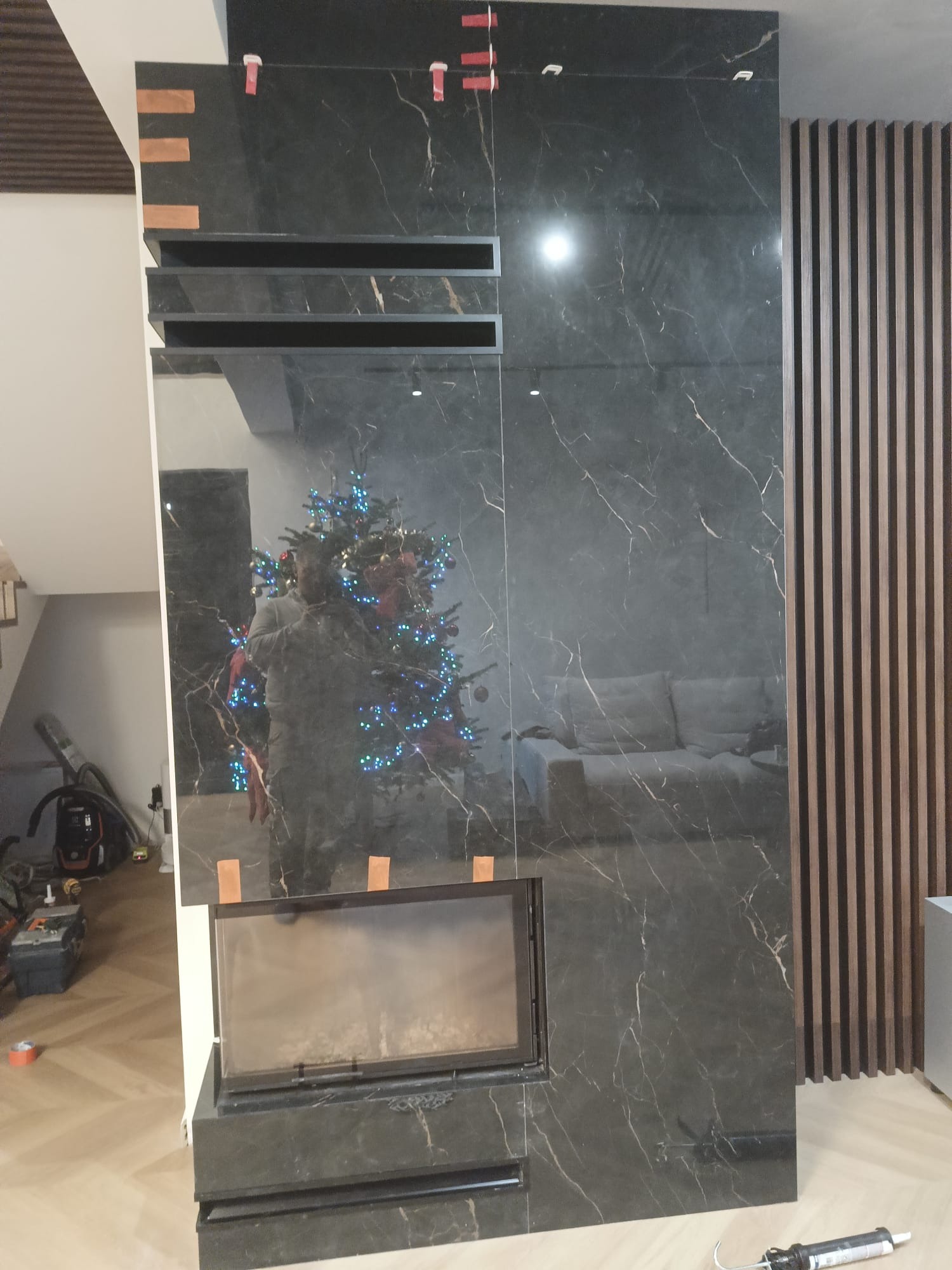 Wykończony kominek z czarnego marmuru z białymi żyłami, obok drewniana ściana z listew. W tle odbicie choinki i sofy. Widoczne narzędzia glazurnicze.