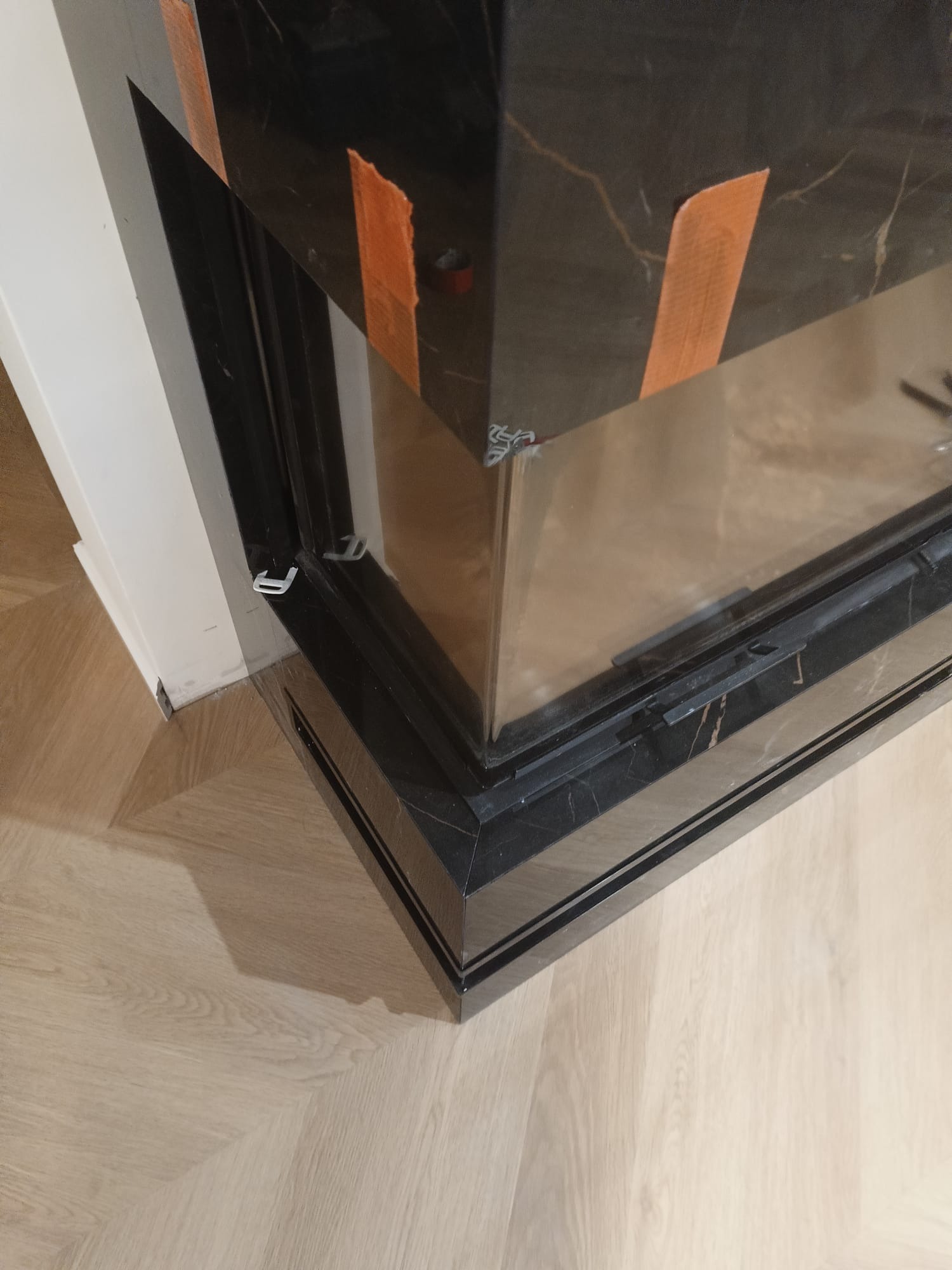 Fragment wykończonego kominka z czarnego marmuru z widocznymi zabezpieczeniami taśmą malarską, na tle parkietu ułożonego w jodełkę i białej ściany.