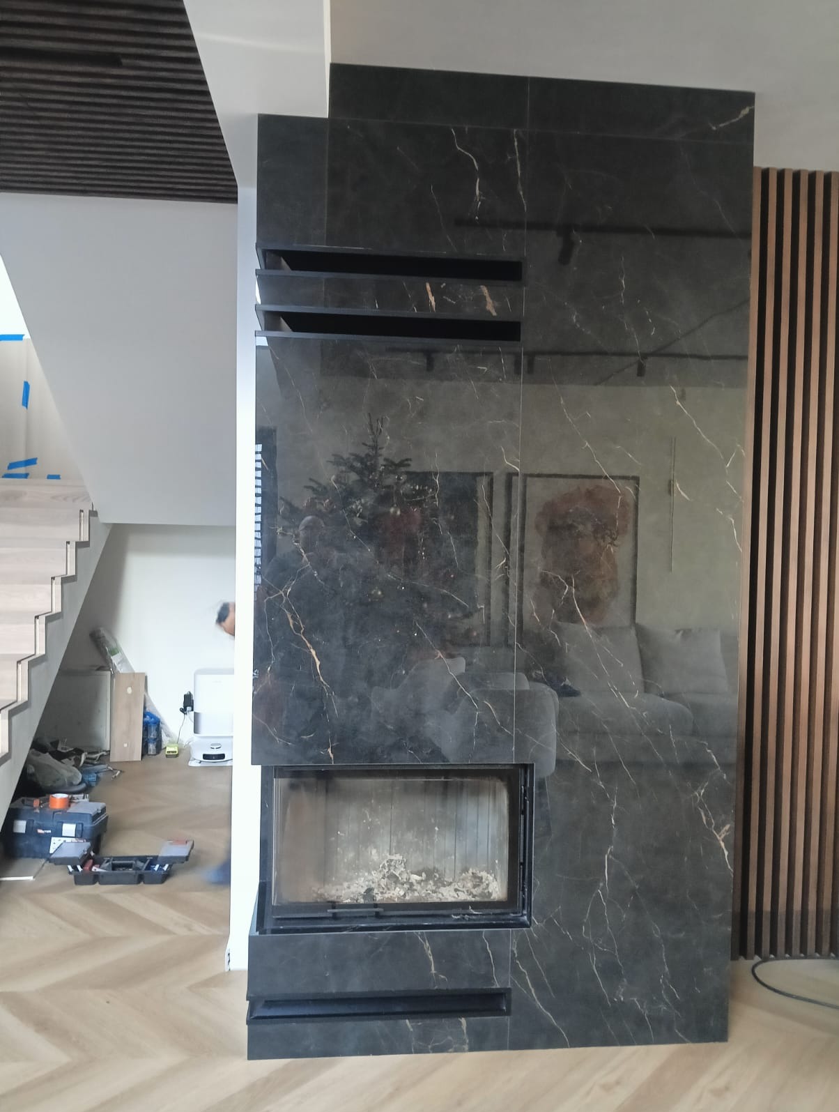 Obudowa kominka z czarnego marmuru z wyraźnym żyłkowaniem, widoczna w salonie z parkietem w jodełkę i schodami. Element dekoracyjny z drewnianych listew po prawej.