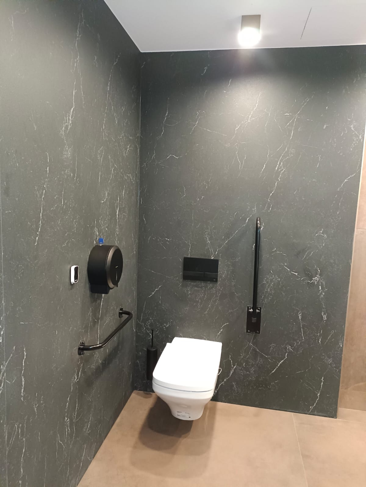 Wykończona łazienka z toaletą, czarnymi płytkami imitującymi marmur, uchwytami dla niepełnosprawnych i dozownikiem mydła. Nowoczesny design.