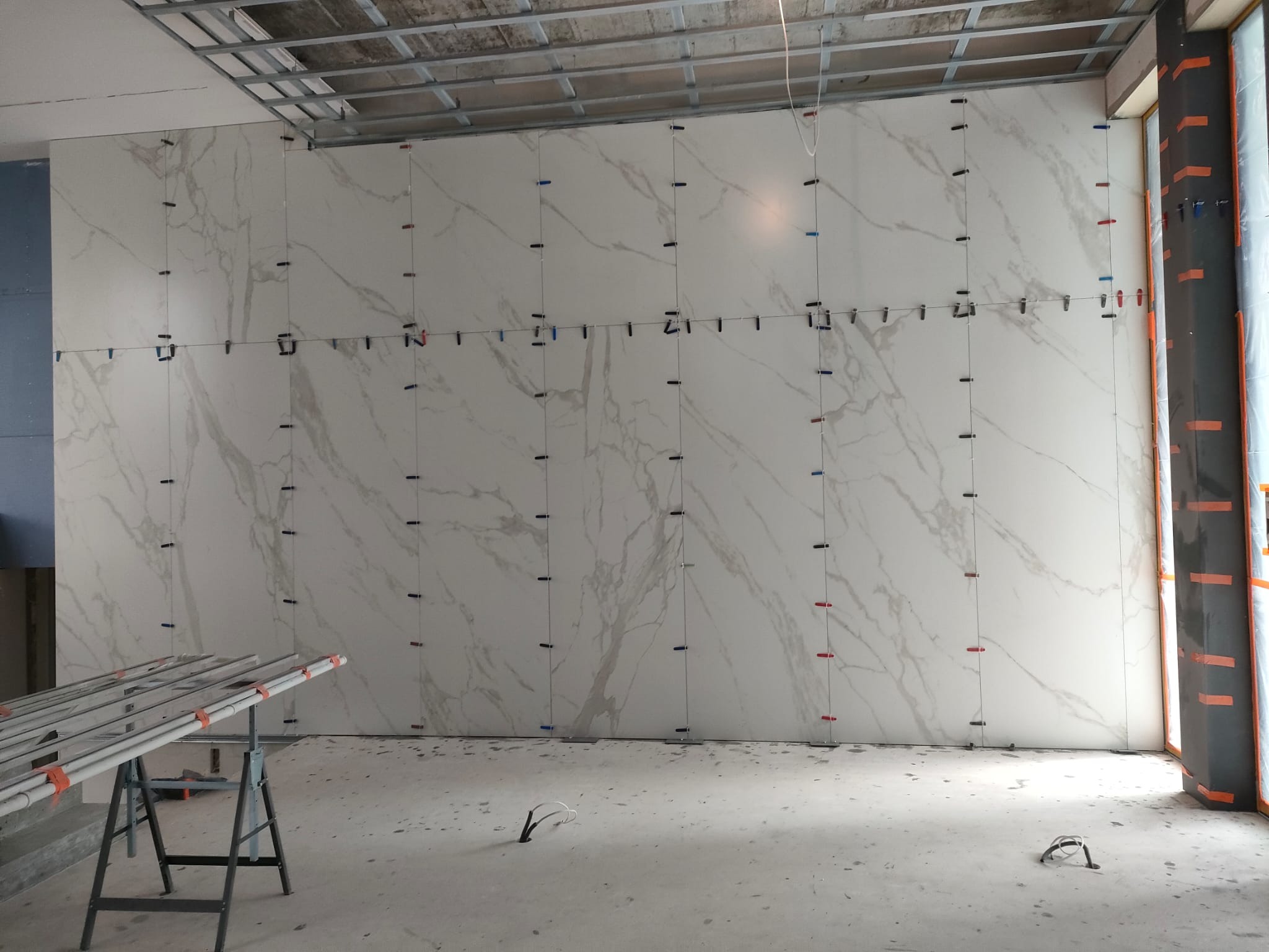 Ściana z białych płytek imitujących marmur, z systemem poziomowania płytek. Widoczne profile sufitowe i zabezpieczone okno. Podłoga surowa, w tle drabina.