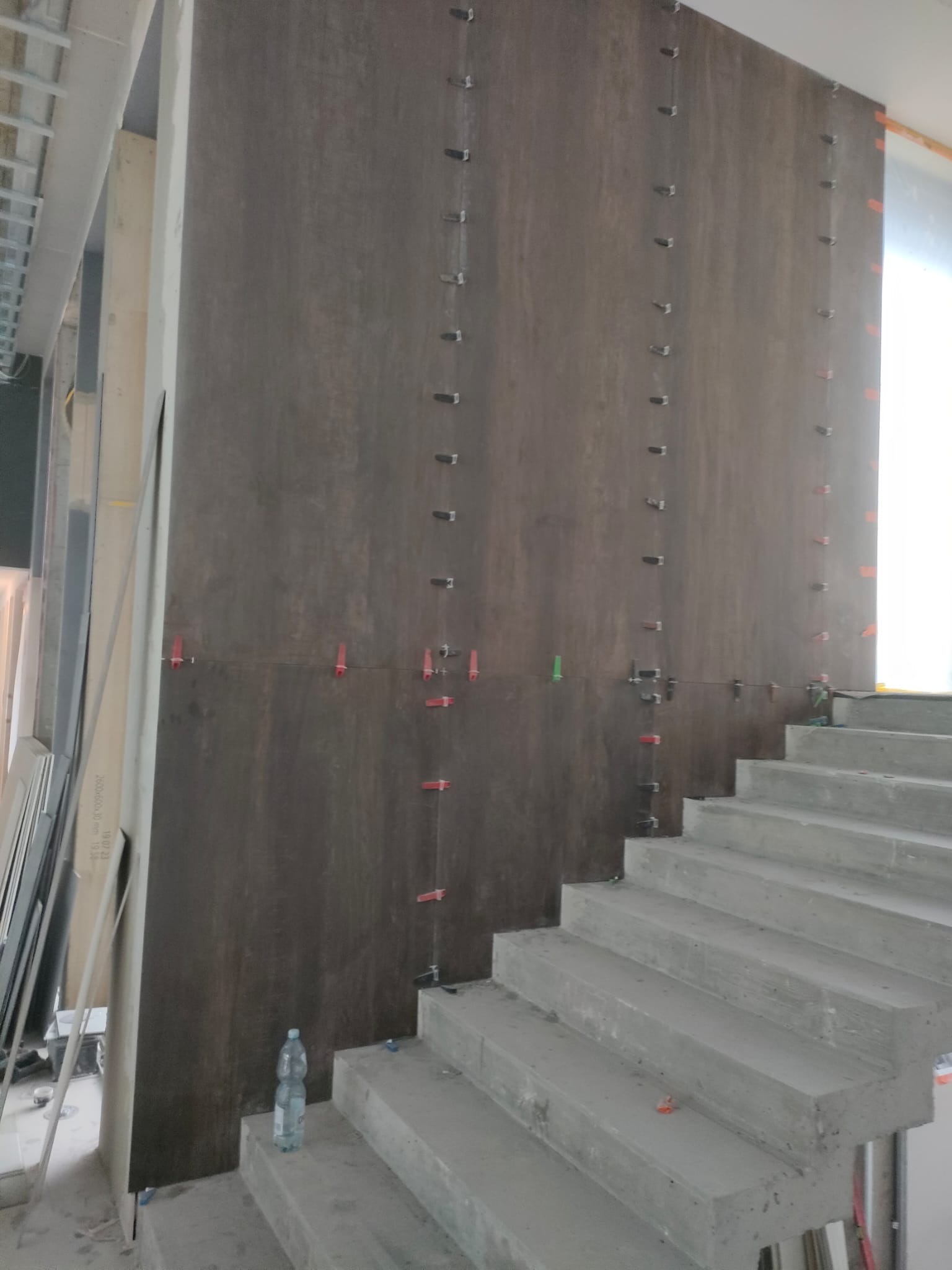 Montaż wielkoformatowych płytek na ścianie przy schodach, z użyciem klipsów poziomujących. Widoczna butelka wody na schodach i płyty w tle.