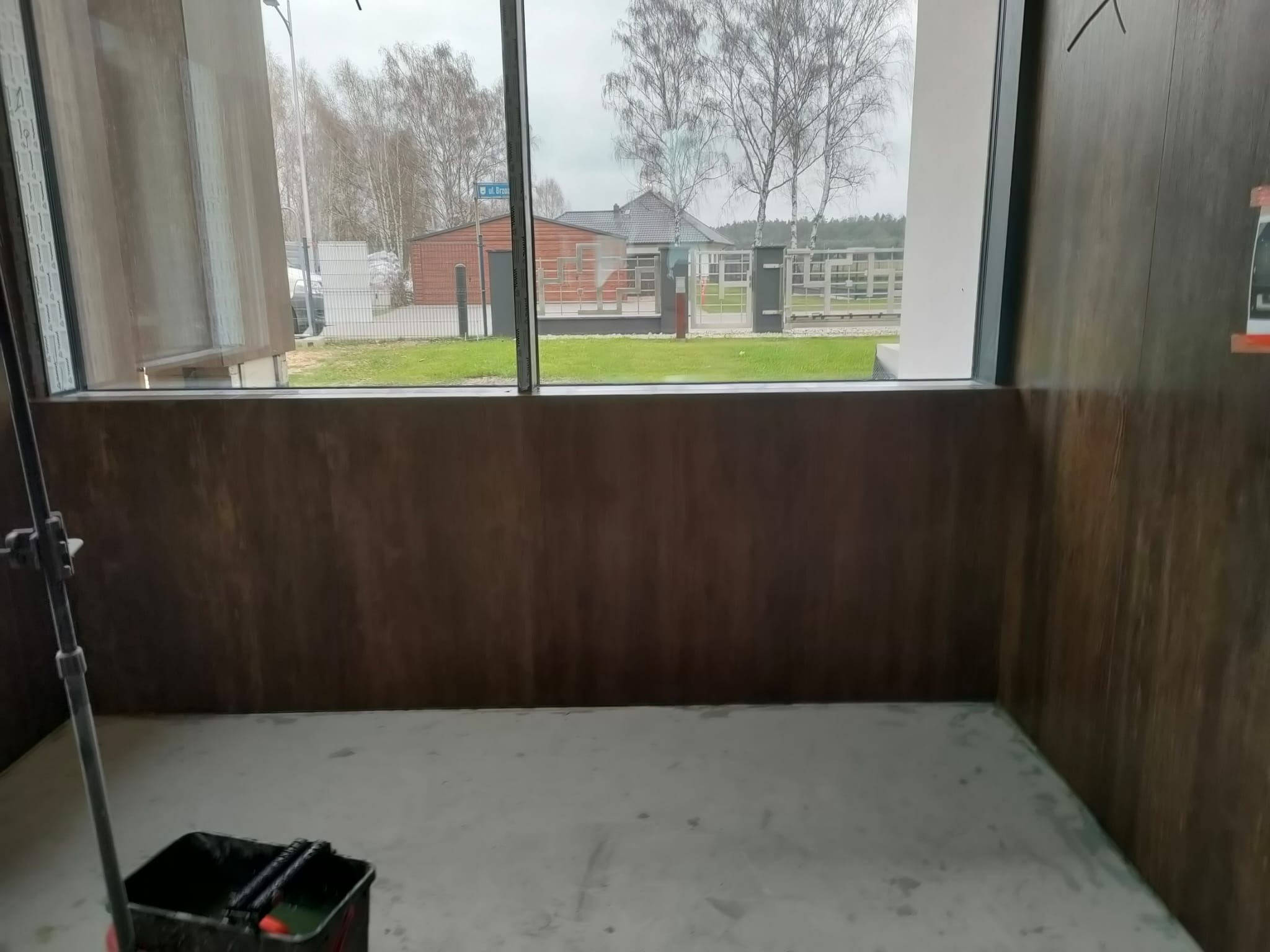 Przygotowane do malowania wnętrze z brązowymi panelami ściennymi i widokiem na zewnątrz przez duże okno. Wiadro z wałkami malarskimi w rogu.