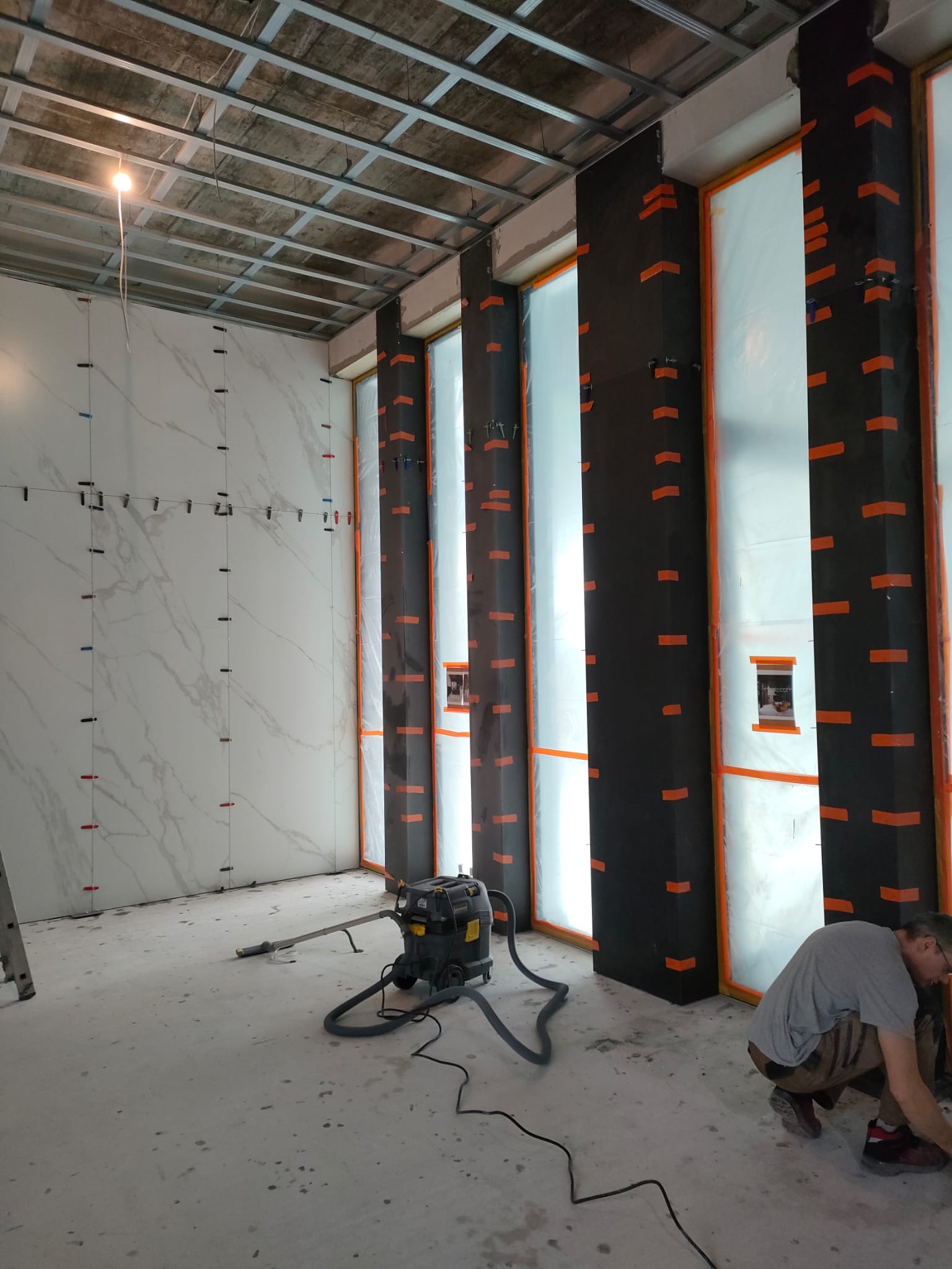 Wnętrze w trakcie prac glazurniczych: ściana z białych płytek imitujących marmur, czarne kolumny zabezpieczone folią, widoczny pracownik i odkurzacz przemysłowy.