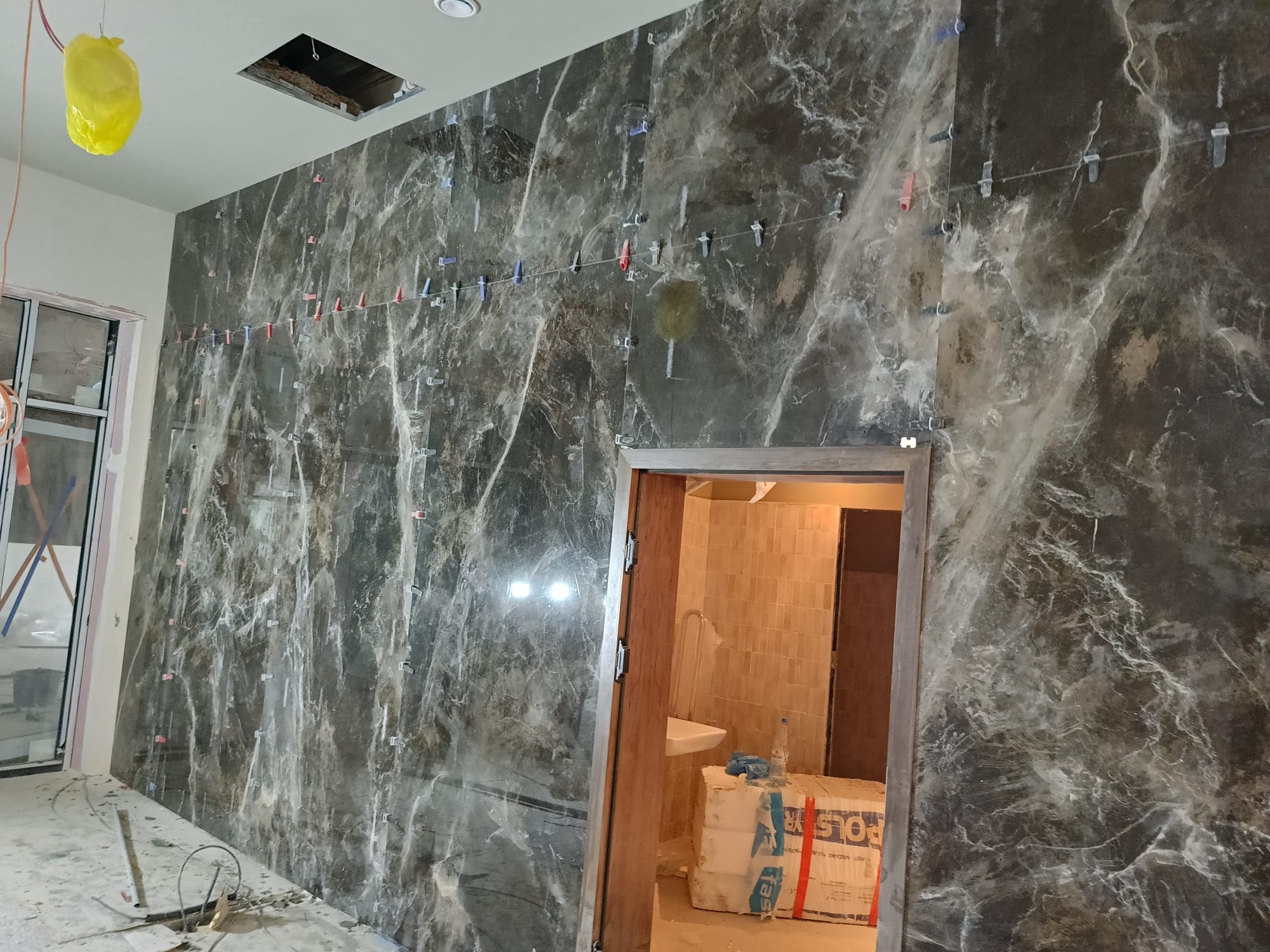 Montaż płytek imitujących marmur na ścianie z użyciem klipsów poziomujących. Widoczny fragment łazienki z umywalką i materiały budowlane.