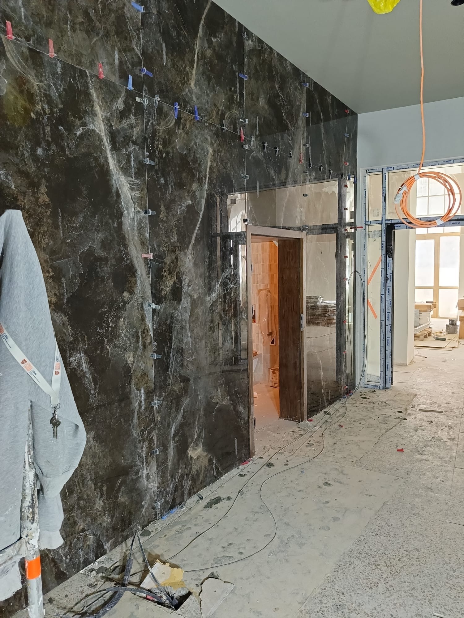 Montaż płytek imitujących marmur na ścianie z użyciem klipsów poziomujących. Widoczne przewody elektryczne i fragment pomieszczenia w trakcie remontu.