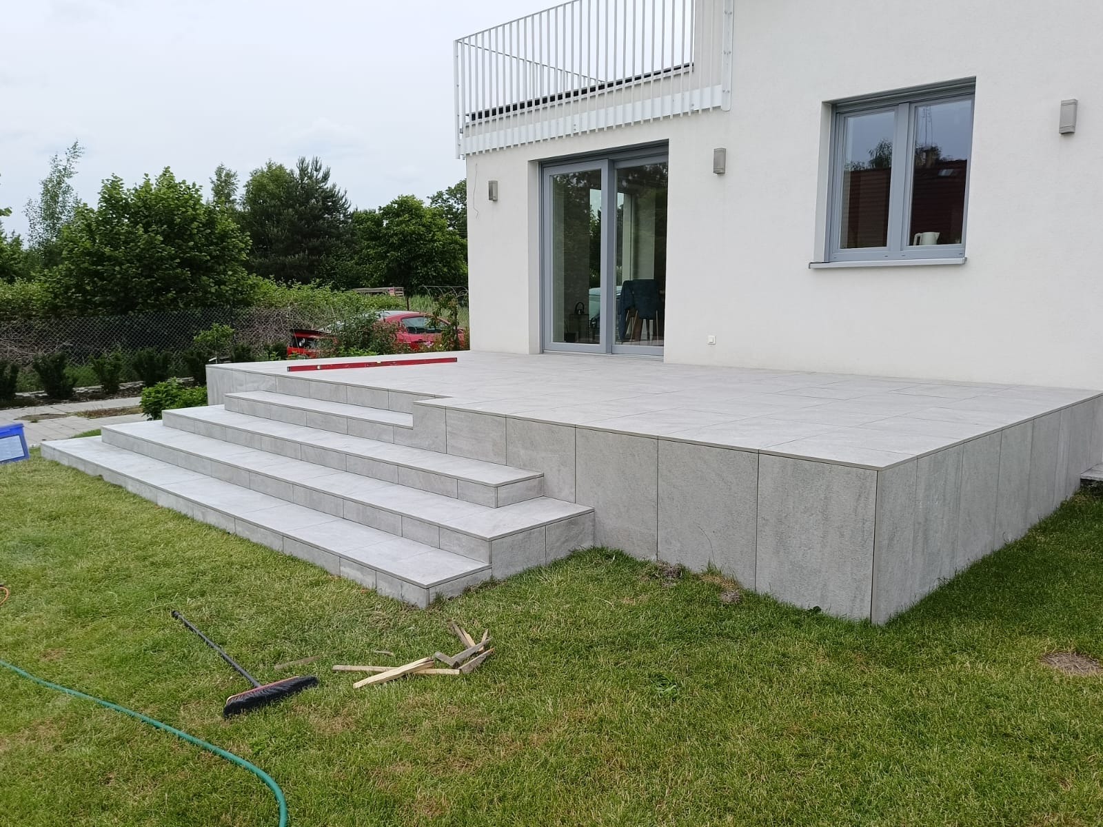 Wykończony taras z szarych płytek ceramicznych, schody z płytek, biały budynek z balkonem w tle, trawnik z narzędziami po lewej stronie.