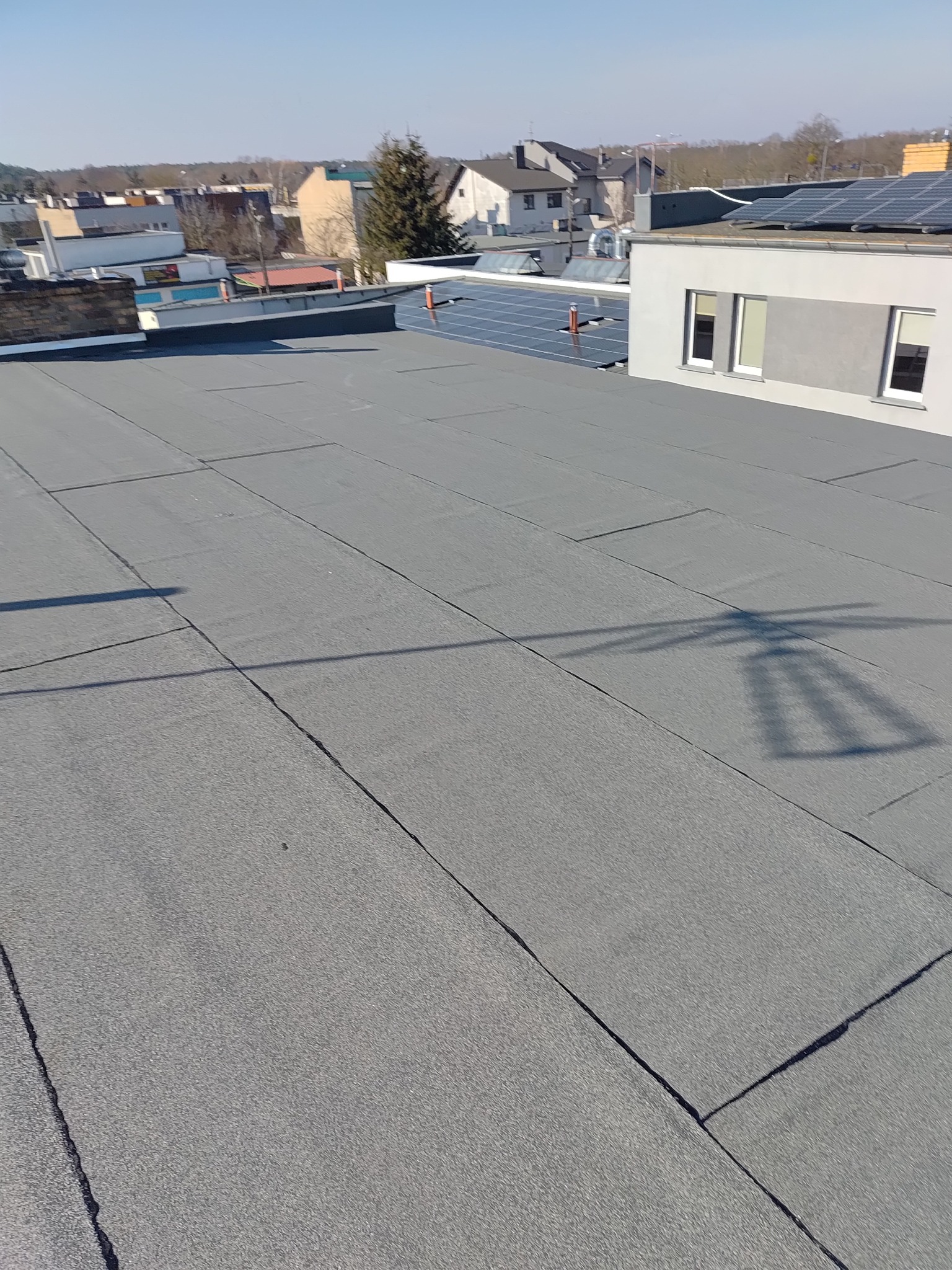 Szary, płaski dach pokryty papą termozgrzewalną w słoneczny dzień. Widoczne panele słoneczne na sąsiednim budynku i zabudowa miejska w tle.