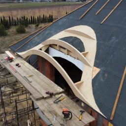 ACC Rafter Alan Piwowar - Wysoki kąt: Dach z papy bitumicznej z drewnianą konstrukcją okna dachowego. Rusztowanie z narzędziami obok muru. W tle drzewa i pole.