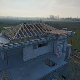 ACC Rafter Alan Piwowar - Budowa domu w trakcie: wzniesione ściany z betonu komórkowego, częściowo wykonana drewniana konstrukcja dachu. Widok z góry na budynek i okolicę.