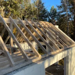 Jasna, drewniana konstrukcja dachu w trakcie budowy, widoczna na tle zielonych drzew i błękitnego nieba. Elementy więźby dachowej ułożone regularnie.