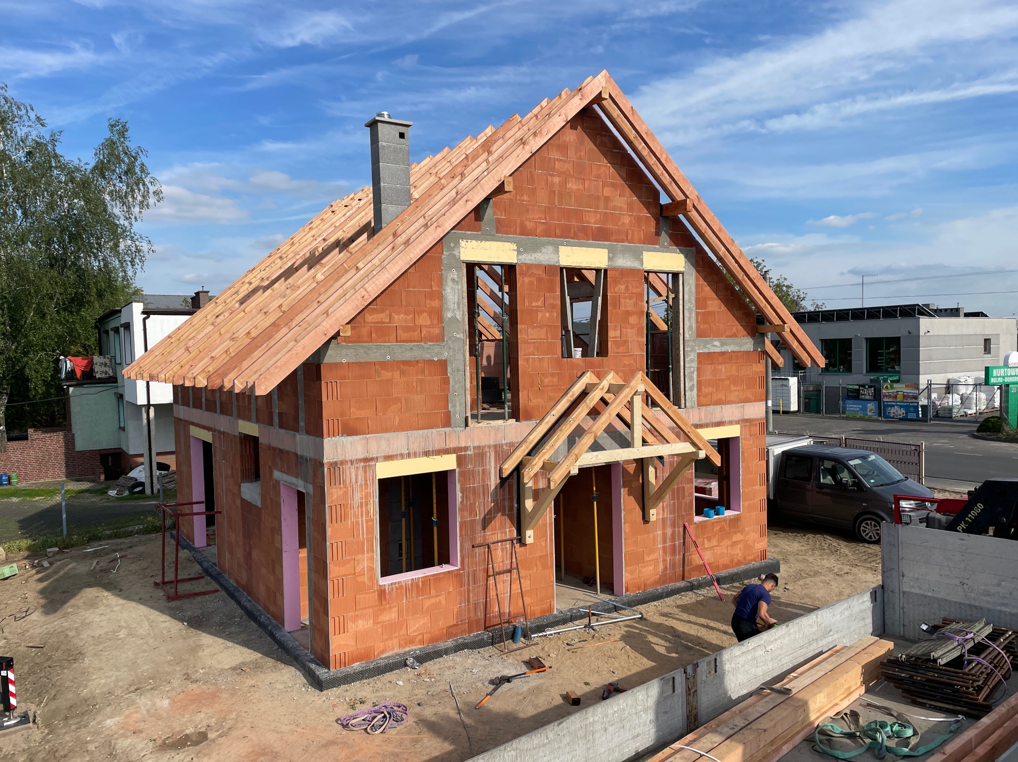 Budowa domu z czerwonej cegły w Sieradzu, widoczna więźba dachowa i komin. Na pierwszym planie pracownik budowlany. Błękitne niebo w tle.