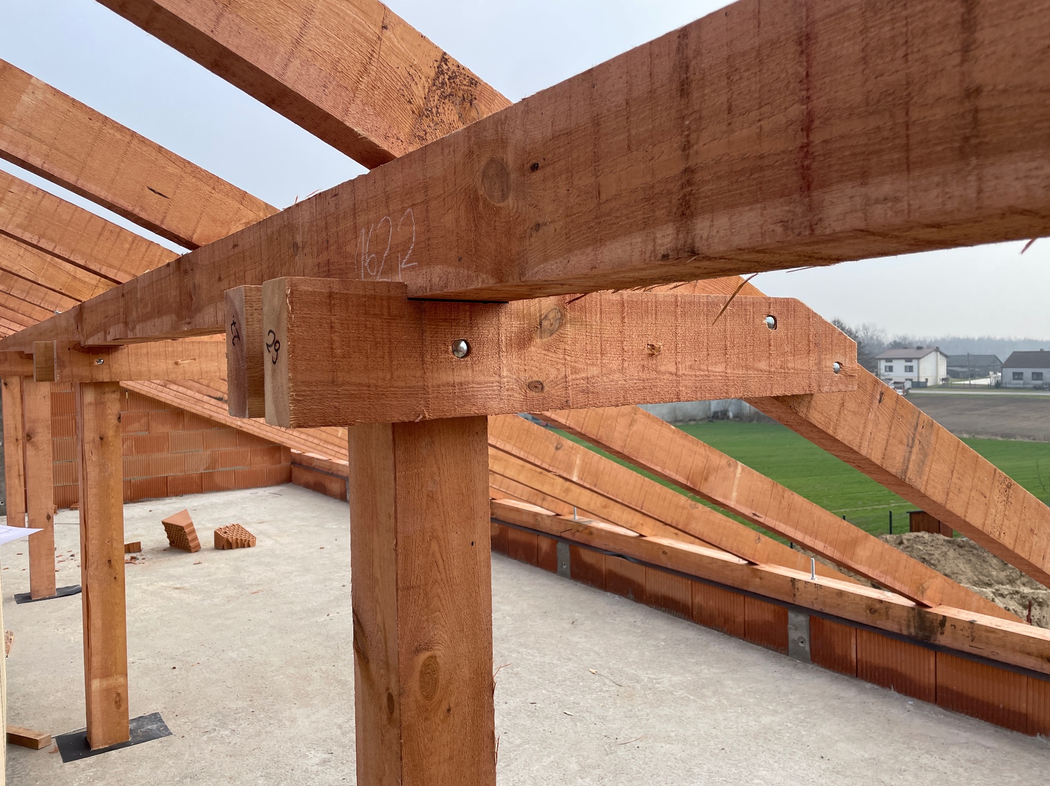 Konstrukcja drewniana dachu w trakcie montażu, widoczne elementy więźby z numeracją, tło stanowią domy i zielone pola.