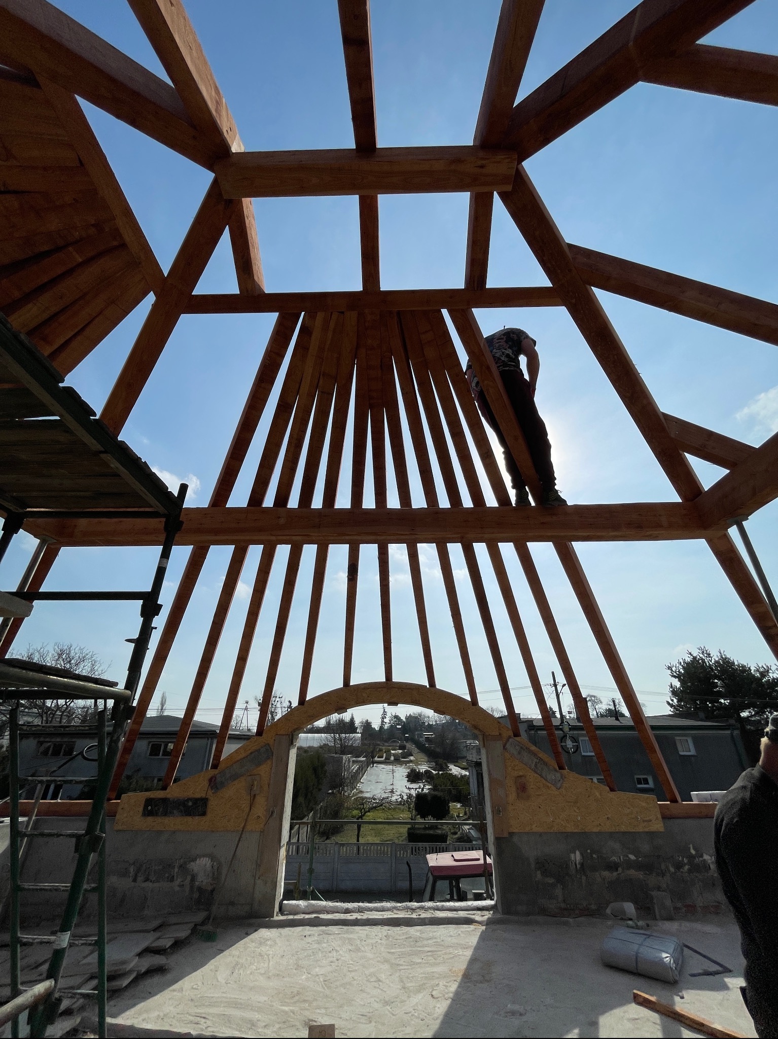 Konstrukcja drewniana dachu w trakcie budowy, widok z dołu na niebo i sylwetkę pracownika. Elementy konstrukcyjne tworzą geometryczny wzór.
