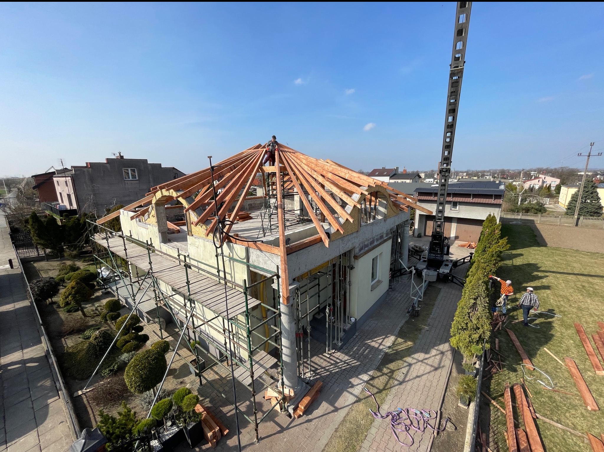 Budowa dachu z drewnianą konstrukcją, widok z góry na dom w trakcie budowy. Na dachu pracuje dekarz, widoczne rusztowania i dźwig. Słoneczny dzień.