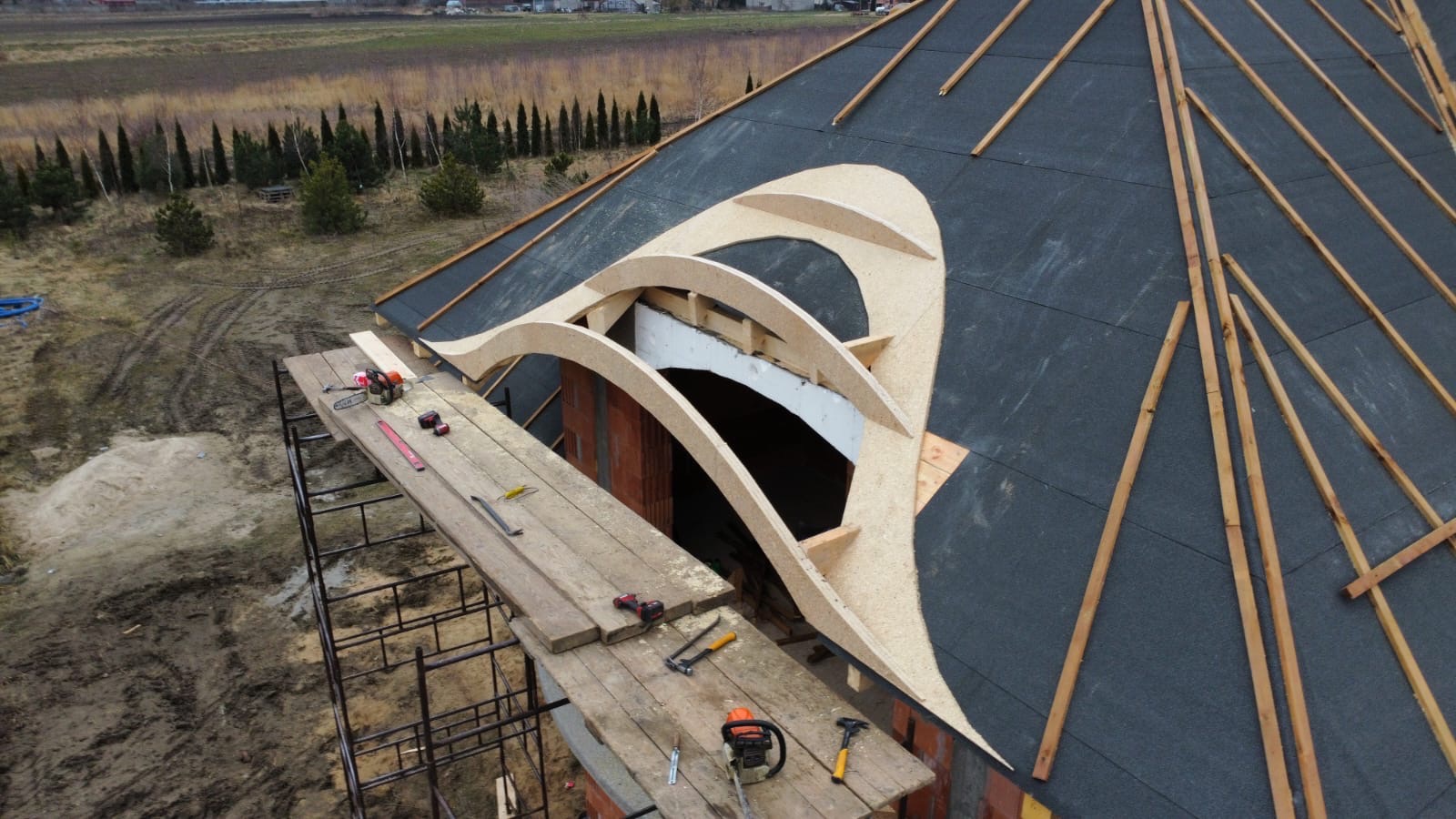 Wysoki kąt: Dach z papy bitumicznej z drewnianą konstrukcją okna dachowego. Rusztowanie z narzędziami obok muru. W tle drzewa i pole.