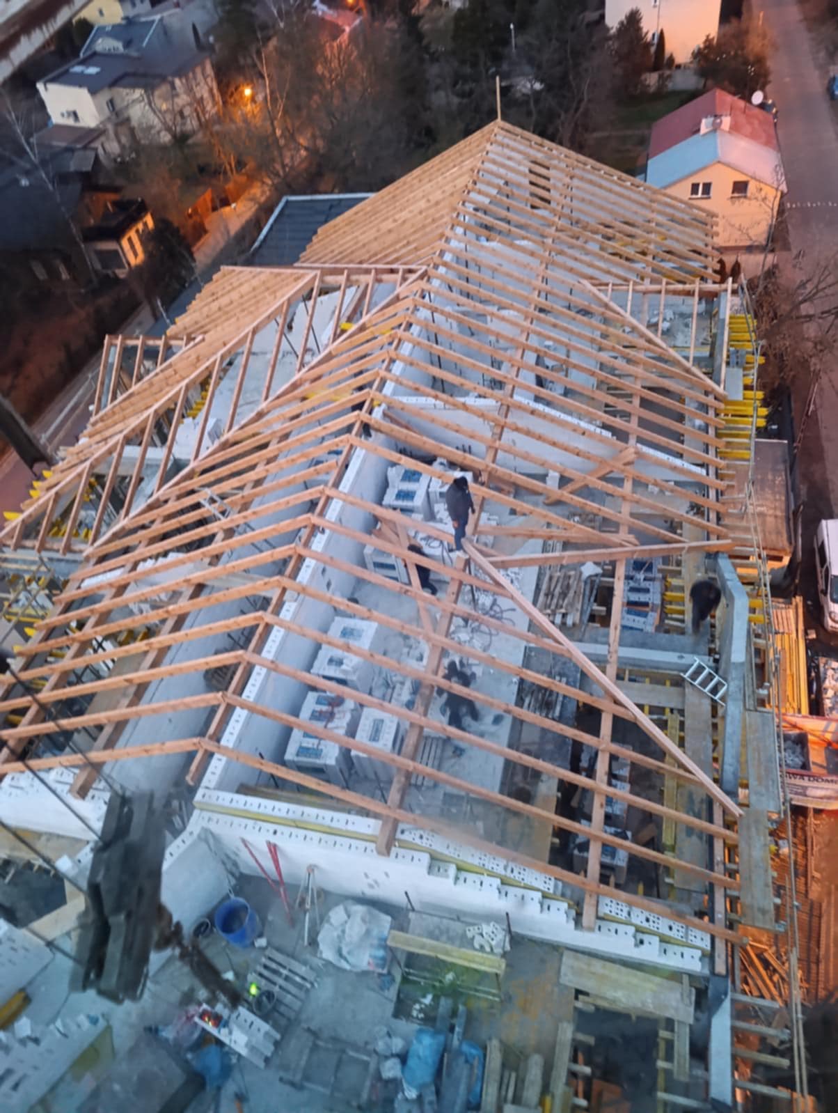 Montaż drewnianej konstrukcji dachu na nowym budynku z widocznymi pracownikami. Ujęcie z góry ukazuje postęp prac w świetle dziennym.