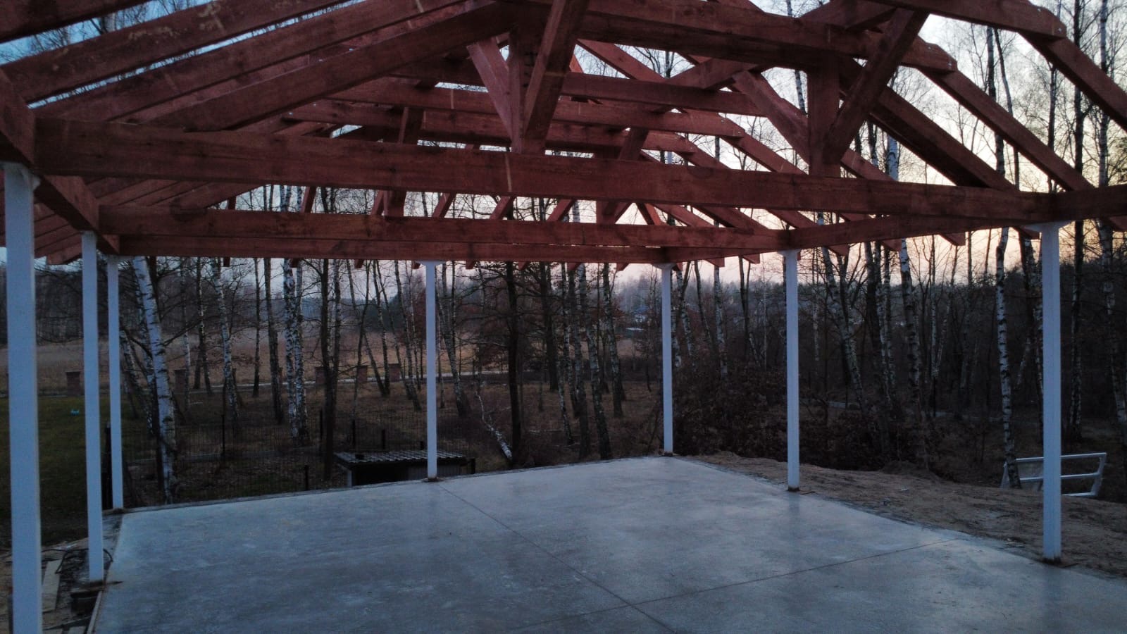 Konstrukcja drewniana z białymi słupami na betonowej płycie, widok na las brzozowy. Elementy konstrukcji w kolorze ciemnego drewna.