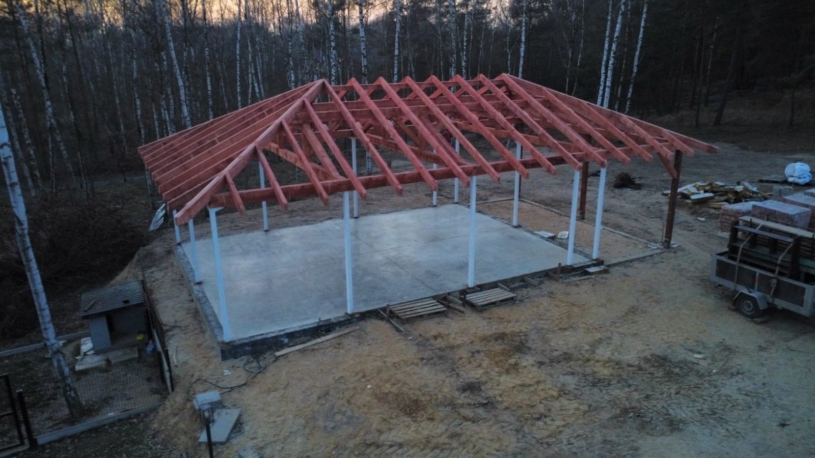 Wznoszenie szkieletu dachu z czerwonego drewna na betonowej płycie fundamentowej. Widoczne białe słupy nośne i las brzozowy w tle.