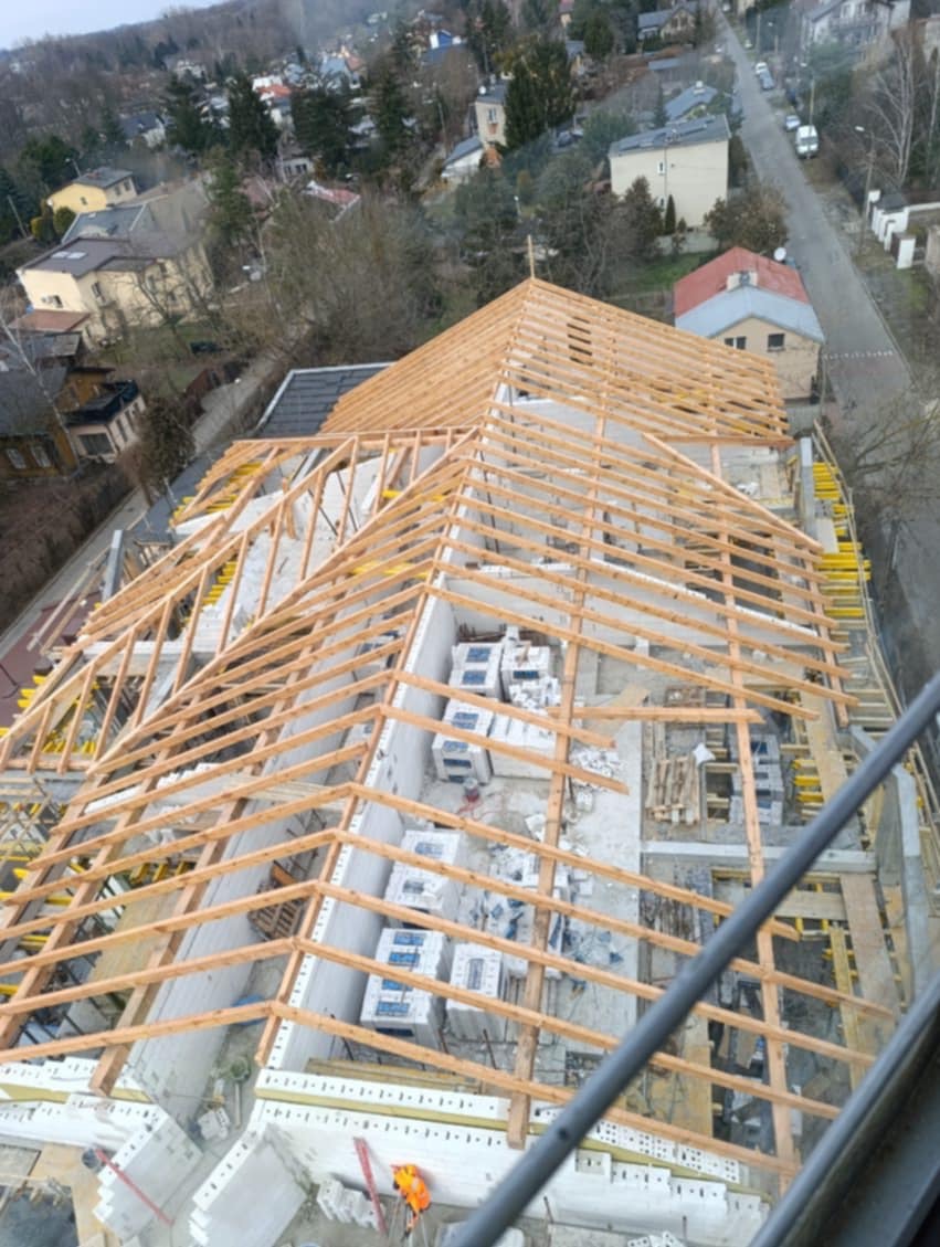 Budowa dachu z drewnianą konstrukcją, widok z góry na budynek w trakcie wznoszenia. Elementy szalunkowe i pustaki na ścianach. Ujęcie z wysokości.