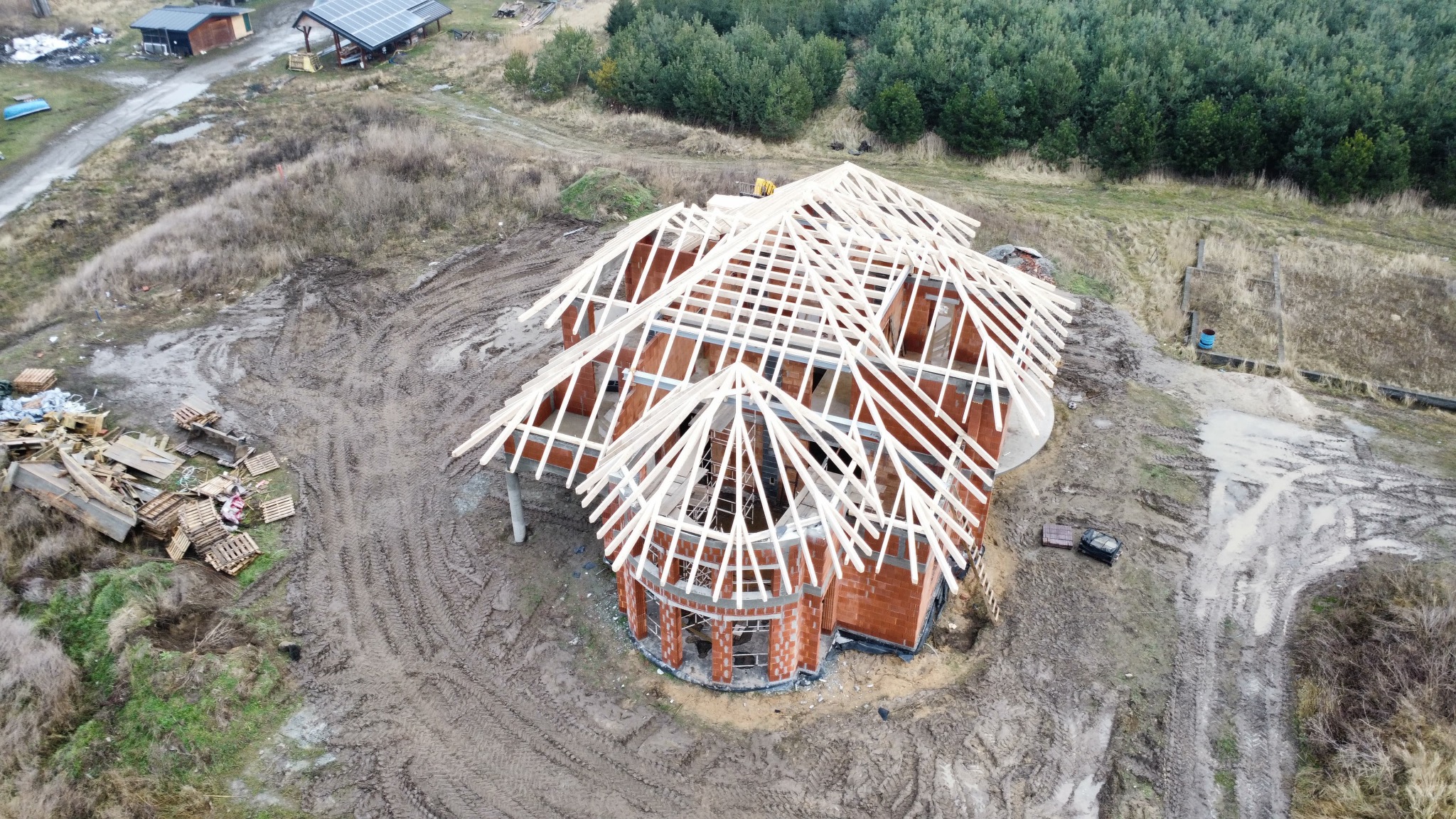 Budowa domu z cegły z drewnianą konstrukcją dachu w surowym otoczeniu, widok z góry. Na pierwszym planie palety i gruz. W tle las.