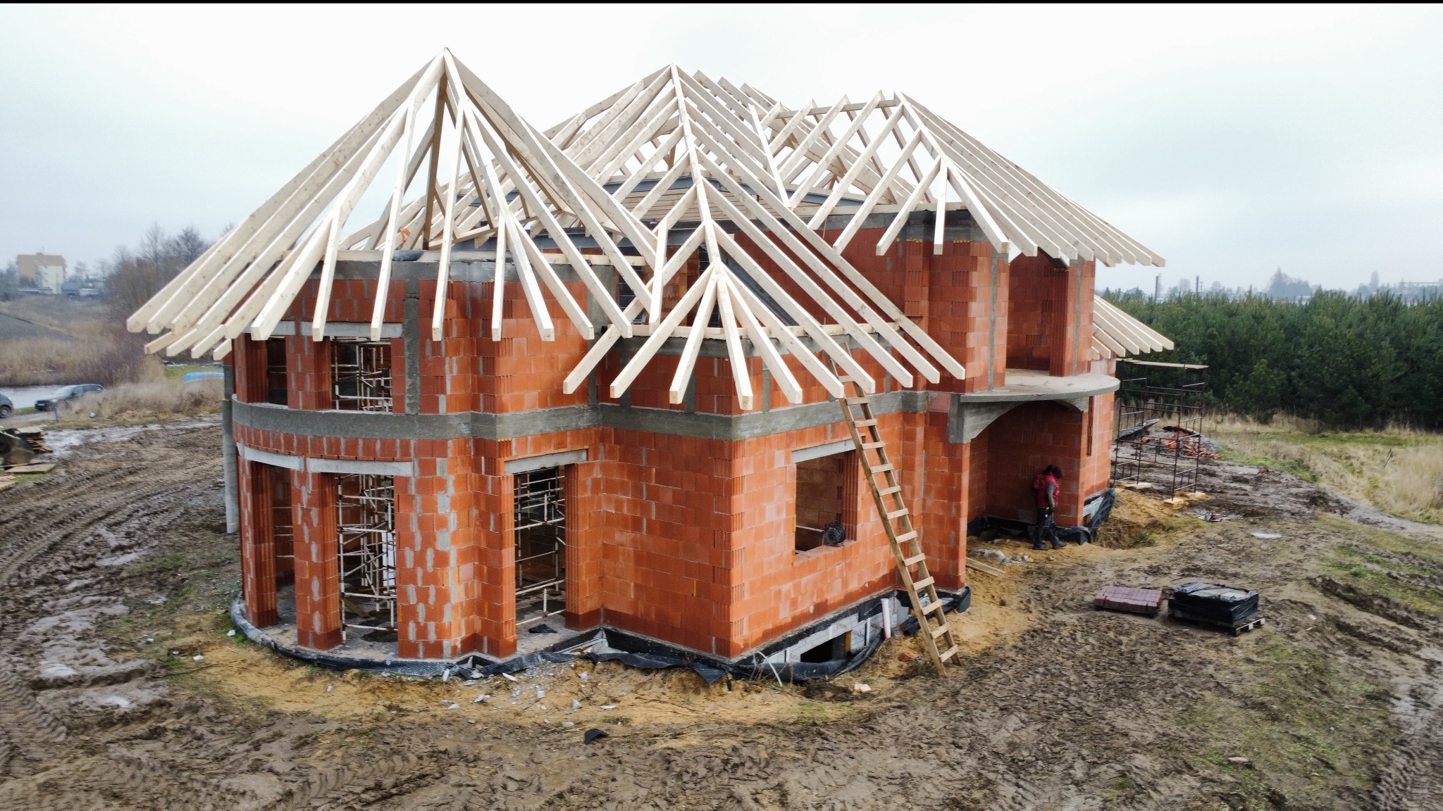 Budowa domu jednorodzinnego: murowane ściany z pustaków i drewniana konstrukcja dachu w trakcie montażu. Na ziemi błoto, w tle las.