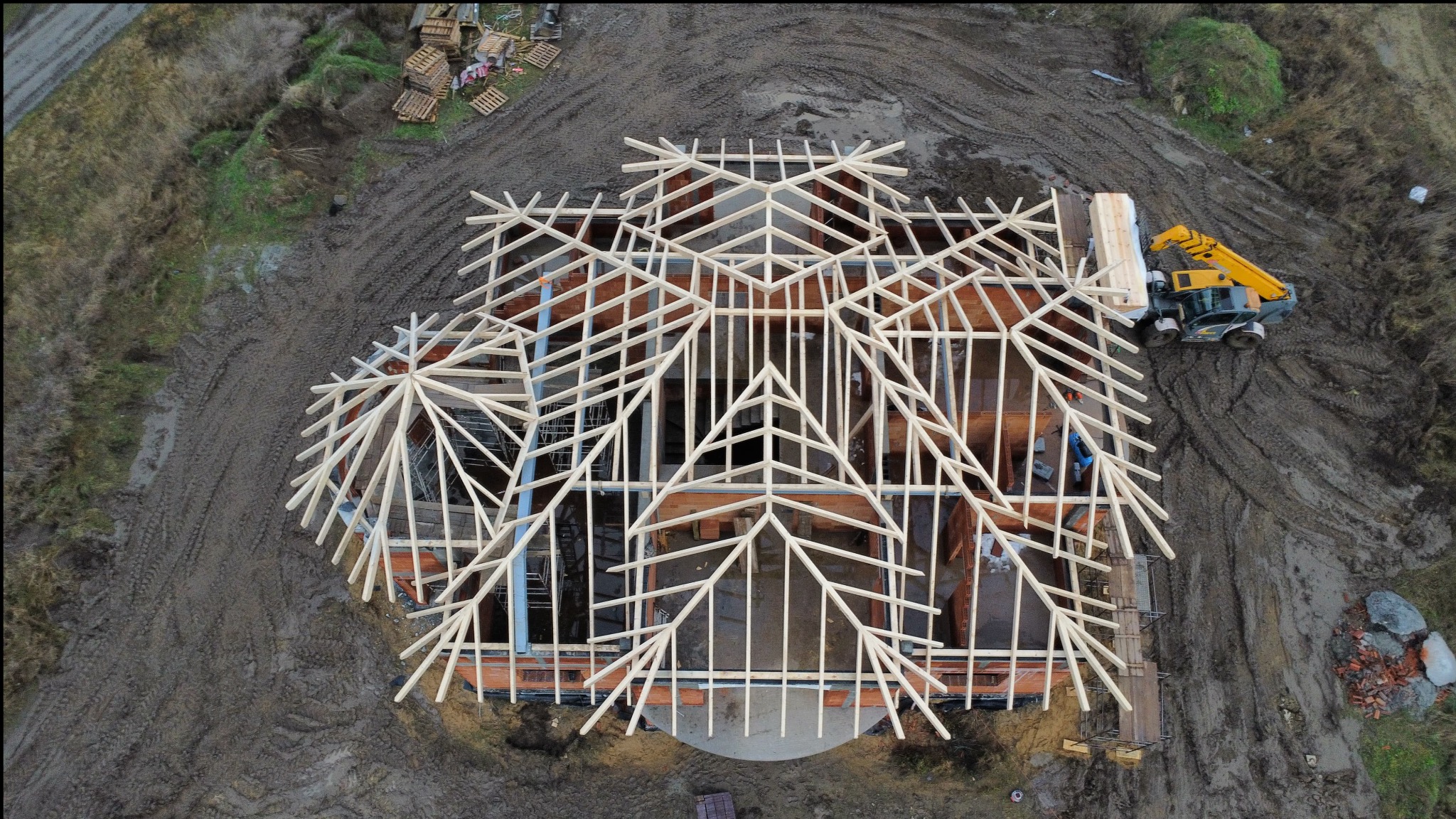 Widok z góry na konstrukcję drewnianą dachu domu w trakcie budowy, z żółtą maszyną budowlaną obok. Teren wokół rozkopany, błotnisty.