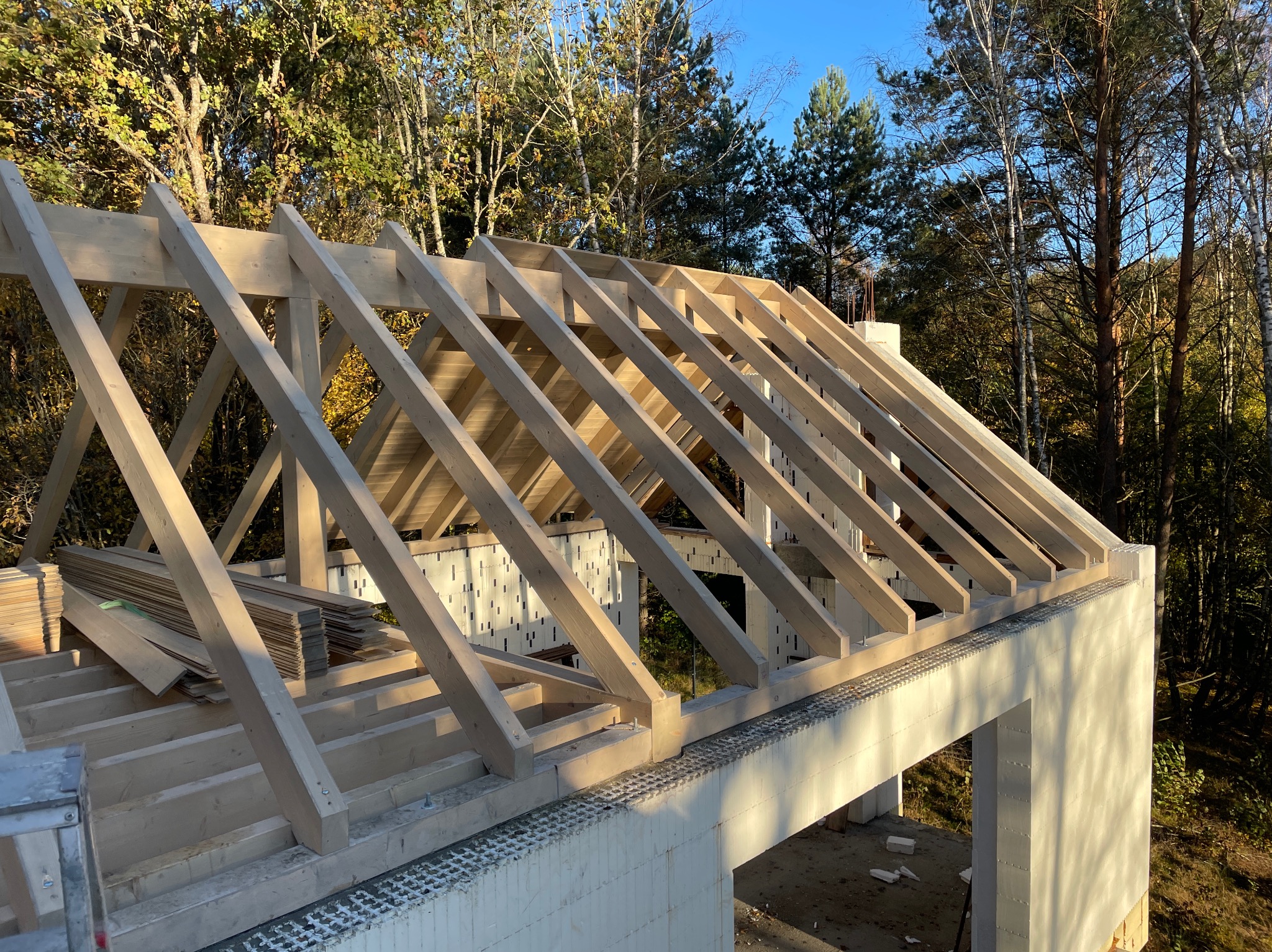 Jasna, drewniana konstrukcja dachu w trakcie budowy, widoczna na tle zielonych drzew i błękitnego nieba. Elementy więźby dachowej ułożone regularnie.