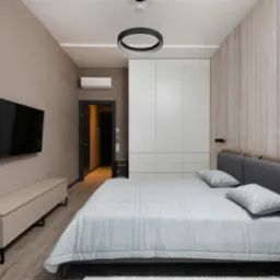 Nowoczesna sypialnia z klimatyzatorem nad drzwiami, minimalistycznym oświetleniem i drewnianą ścianą. Telewizor wisi na ścianie obok szafki RTV.