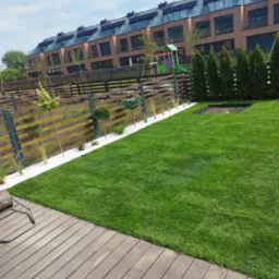 Zadbany trawnik z rolki przylegający do drewnianego tarasu, z widokiem na nowoczesne szeregowce i ogrodzenie z białym kamieniem dekoracyjnym.