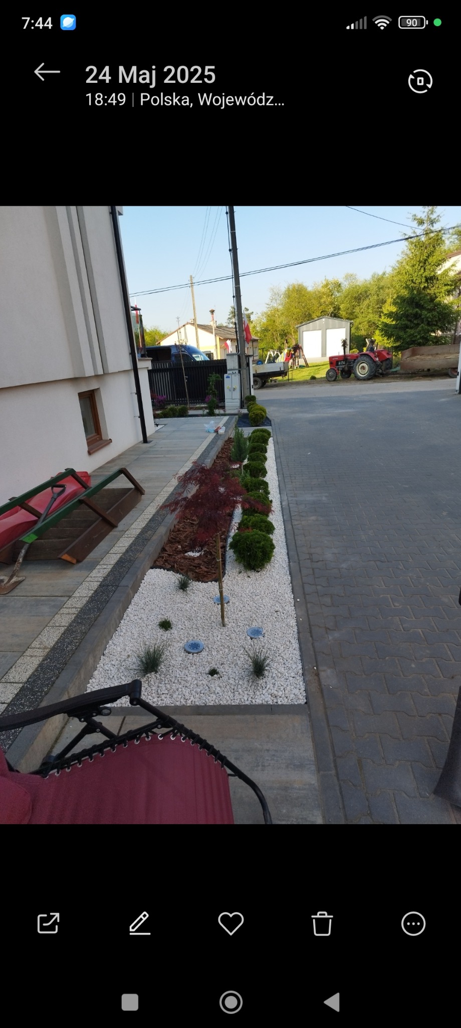 Nowoczesny ogród z białym żwirem, ozdobnymi krzewami i klonem palmowym wzdłuż ścieżki, oświetlony lampami gruntowymi. W tle traktor i budynek.