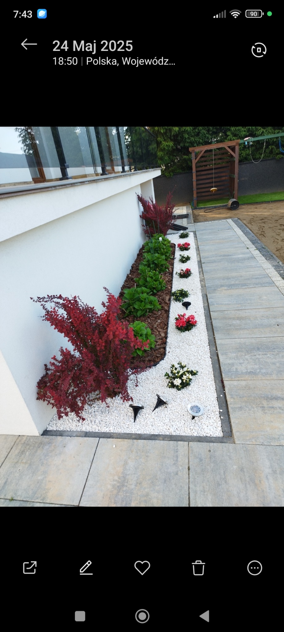 Rabata z białym grysem, ozdobnymi krzewami i kwiatami przy białym budynku. Ścieżka z płyt betonowych. Oświetlenie rabaty lampami solarnymi. Huśtawka ogrodowa w tle.