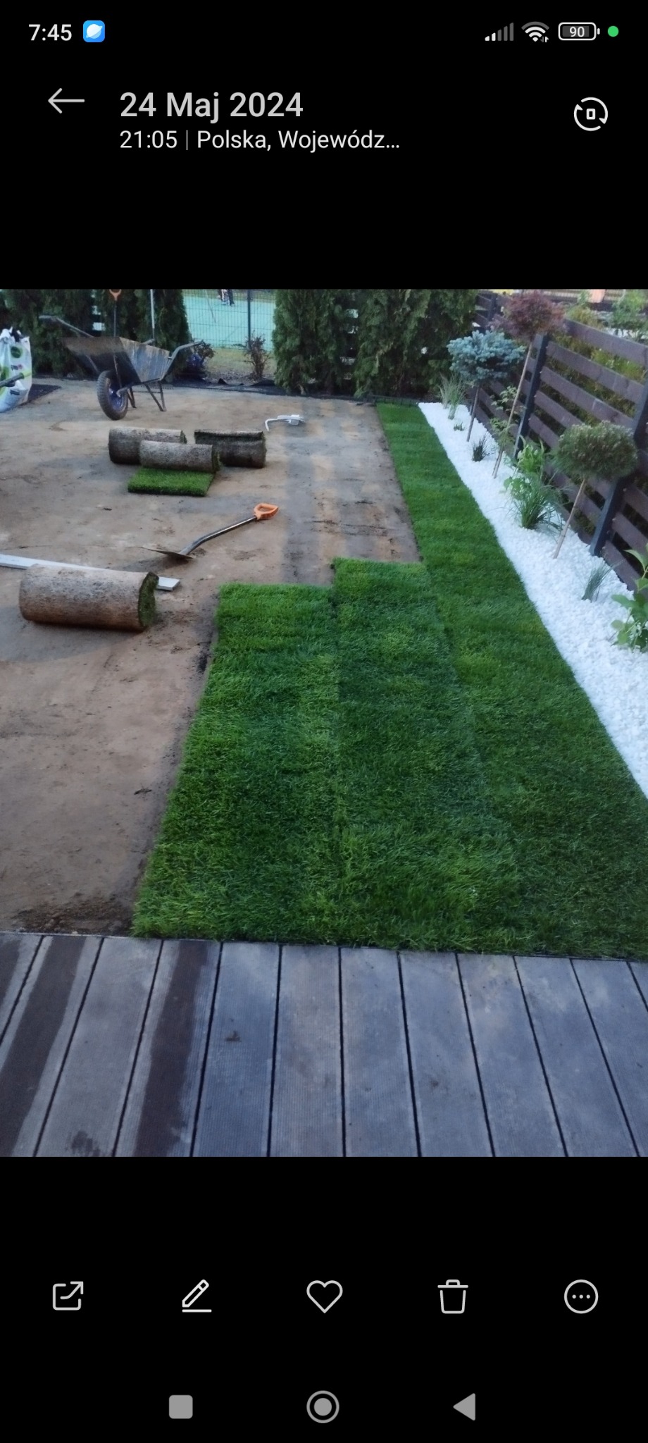 Układanie trawy z rolki na przygotowanym terenie. Widoczne rolki trawy, łopata, taczka i białe kamienie dekoracyjne wzdłuż ogrodzenia. Częściowo ułożona trawa przy drewnianym tarasie.