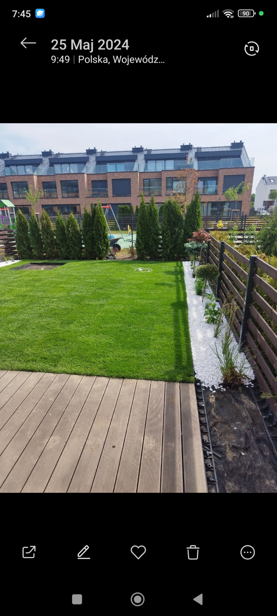 Zadbany ogród z równo przystrzyżonym trawnikiem, tujami i drewnianym tarasem. Białe kamienie dekoracyjne oddzielają trawnik od rabaty. W tle budynki mieszkalne.